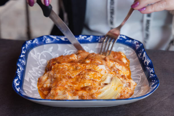 Elegant blue dish holds creamy baked pasta in rich tomato sauce, topped with cheese.  A person enjoys this comforting meal with knife and fork.