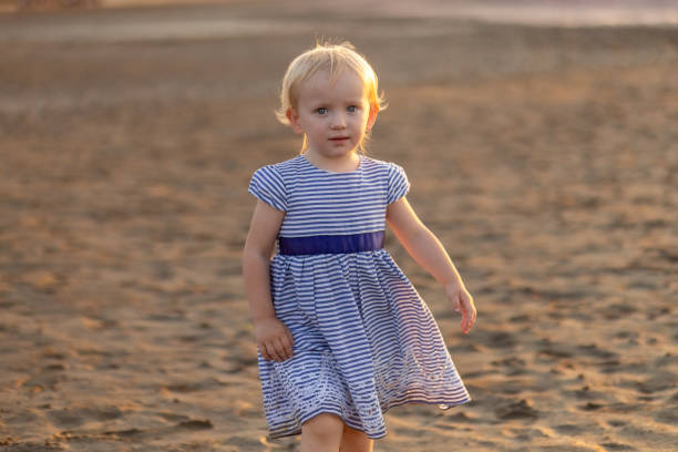 A young child in a striped dress strolls along a sandy beach, the warm light of golden hour illuminating the scene and creating a peaceful atmosphere around her.