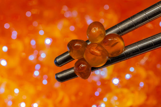 Red caviar is elegantly presented on stainless steel chopsticks, with glowing salmon eggs softly blurring the background