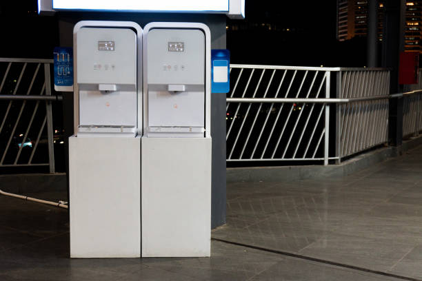 istock Two white water dispensers stand side-by-side on a platform, illuminated in an outdoor night setting. A fence is seen in the background. 2196671478