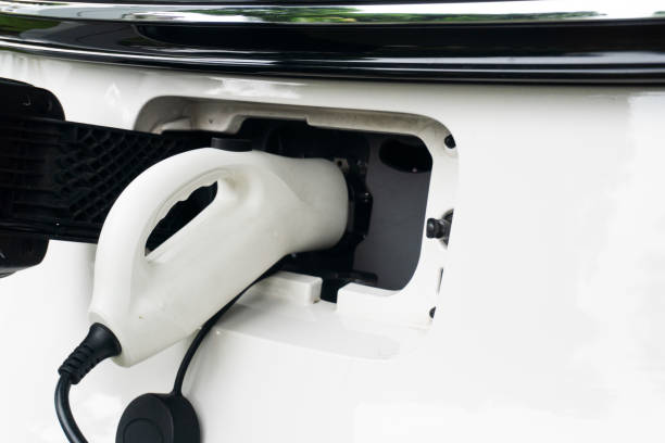 A white electric car is being charged, a white charger inserted into the vehicle's charging port, close up view.