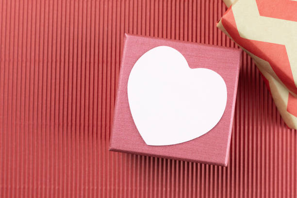 Heart-shaped note paper over red gift box on red corrugated paper background. Concept for Valentine's Day, Mother's Day, and Birthday greeting card. Top view, copy space. Positive emotion.