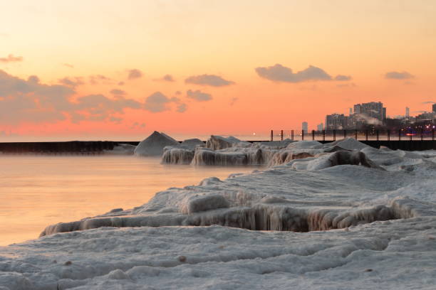 A high-angle drone shot featuring the historic Port Dalhousie Pierhead Lighthouse at the end of a snow-covered pier. The image showcases the frozen shoreline and the icy waters of Lake Ontario in St. Catharines, Ontario. The vibrant green accents of the lighthouse stand out against the white snow and cold blue-green water, capturing the essence of a Canadian winter landscape.
