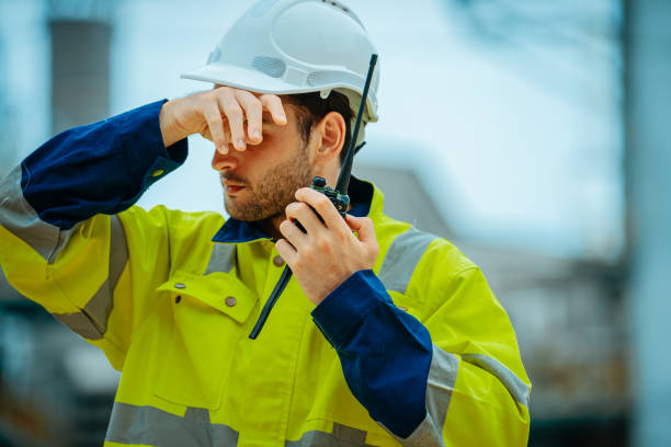 istock Construction worker communicating on radio while shielding eyes from sun at busy worksite during daylight 2196680888
