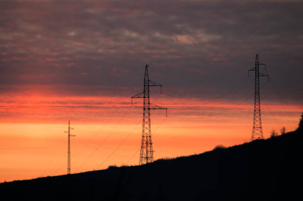 Silhouettes of electricity pylons on a hill against a vibrant sunset sky with shades of orange, red, and purple. The image captures the contrast between industrial structures and the natural beauty of dusk.