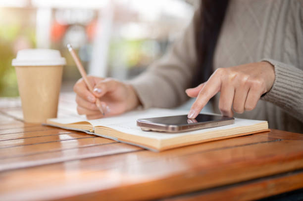 istock A close-up of a woman touching her smartphone screen, checking messages while writing in a book. 2196694088