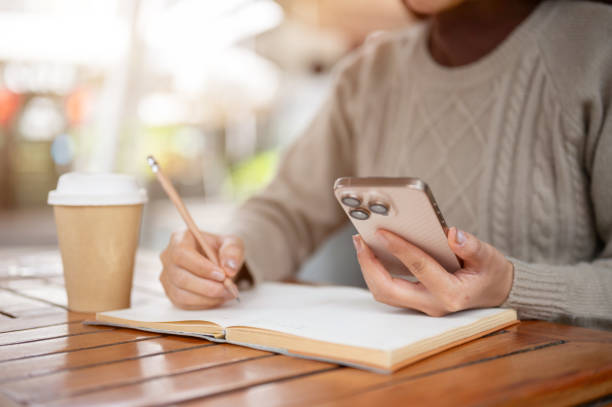 istock A close-up of a woman checking messages on her smartphone while taking notes in a book 2196694097