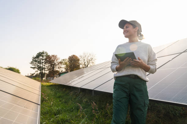 istock Female farmer wearing work uniform stands next to solar panels. Modern agricultural farm using solar panels. Agricultural silos in the background. 2196705552
