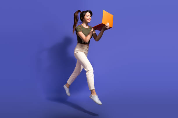 istock Young cheerful woman jumping while using a laptop against a vibrant purple background 2196710740
