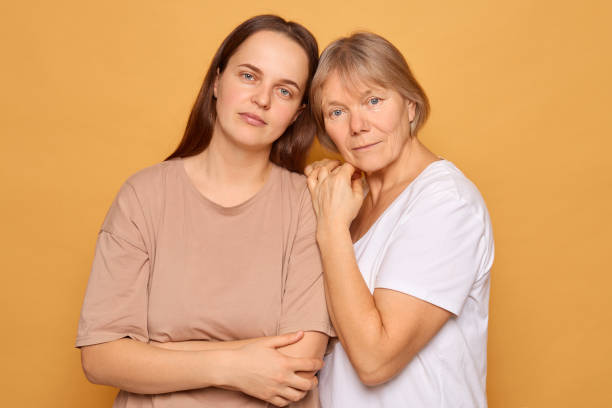A woman stands closely next to her mother, both displaying a deep bond. Their gentle expressions reflect a sense of love and mutual support. The warm yellow backdrop enhances their connection