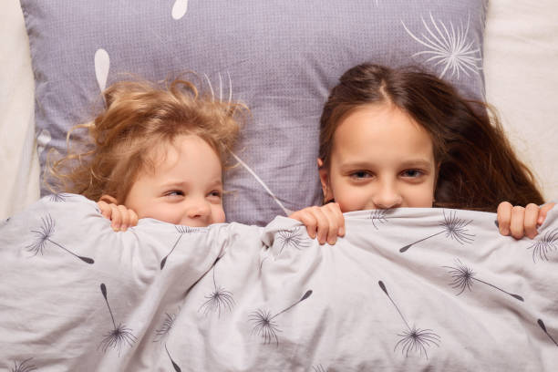 istock Two young sisters share a playful and cozy moment in bed. They smile playfully while peeking out from under the soft blankets, showcasing their close bond and joy in a bright bedroom 2196713471