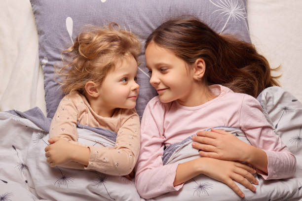 istock Two young sisters lie side by side on a soft bed, sharing laughter and smiles as they snuggle under a cozy blanket in their bedroom during the evening 2196713472