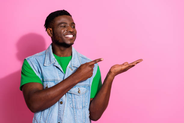 istock Young man in denim vest points cheerfully to side against vibrant pink background, featuring casual fashion and positive energy. 2196715053