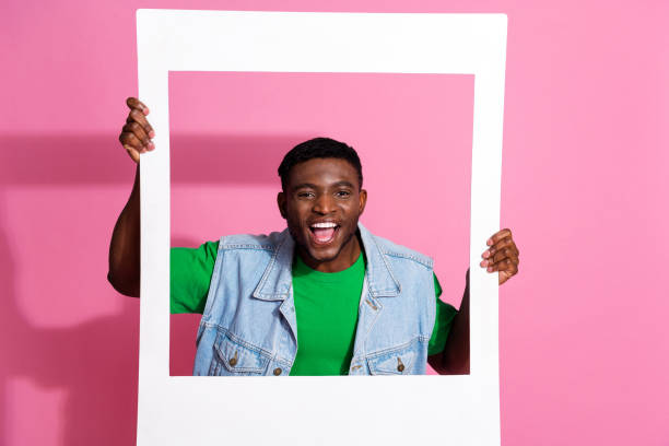 istock Young man in denim vest joyfully posing against a pink background, holding white frame, showcasing cheerful expression. 2196715246