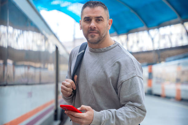 Serious man wearing a gray sweatshirt holding a red smartphone while looking around at a train station. His expression conveys anticipation, travel, and modern connectivity.