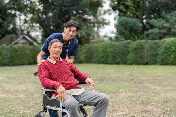 istock Nursing aide proudly standing behind elderly man in wheelchair, showcasing support and companionship in a garden 2196718420