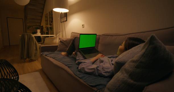 A woman uses a laptop with green screen lying on the couch in an apartment with dim lights, she laughs