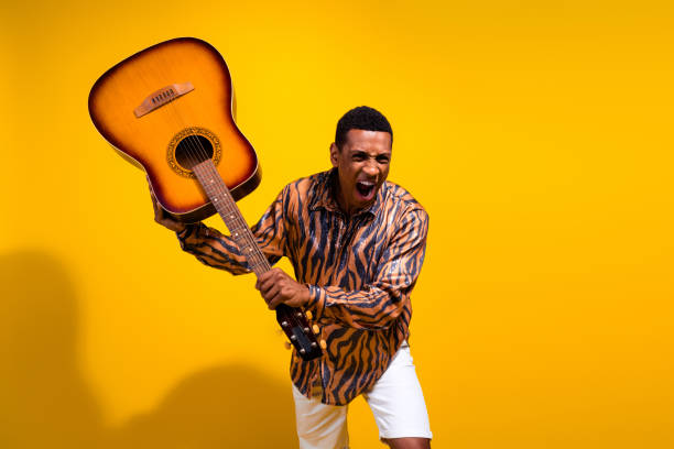Excited young man wearing a stylish patterned shirt, holding a guitar in an expressive pose, set against a vibrant yellow backdrop, conveying energy, fun, and a carefree attitude.
