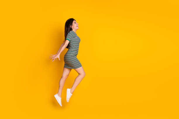 istock Smiling young woman jumps joyfully against a vibrant yellow studio backdrop, dressed stylishly in a striped casual dress and white sneakers, depicting happiness, movement, and a cheerful lifestyle. 2196744290