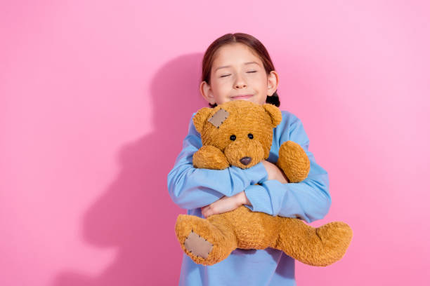 A cheerful preteen girl in a casual blue sweatshirt hugging her brown teddy bear, radiating joy, with a vibrant pink backdrop.