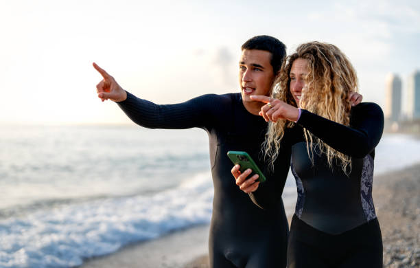 Happy carefree young people, friend, surfers walking on the coast while using mobile phone and enjoying summer vacation and smiling