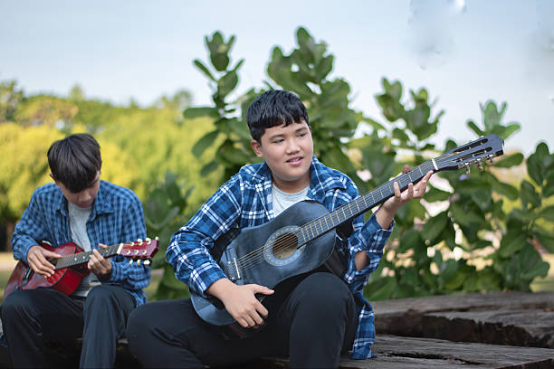 Asian teenaged boy spending his free times with his friend on weekend by practising and playing acoustic guitar or ukulele in the park together, recreational activity of teenagers concept.