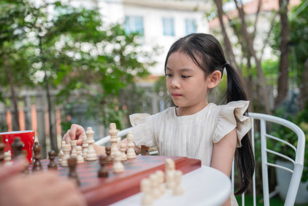 Asian little cute girl thinking over her move in chess during playing challenge, game or play with strategy for competition with her grandfather at home. Asian lovely two generation family play together by board for queen, king and pawn for contest, bonding and retirement
