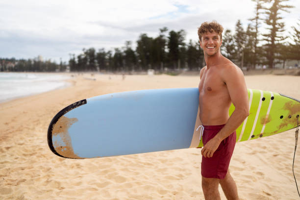 A cheerful surfer standing on Manly Beach, holding a colorful surfboard, enjoying a bright sunny outdoor day near the ocean.