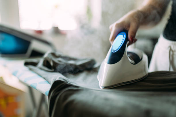Person using a modern steam iron on an ironing board in a well-lit living room, generating visible steam and carrying out the task of ironing clothes, displaying domestic care and household chores