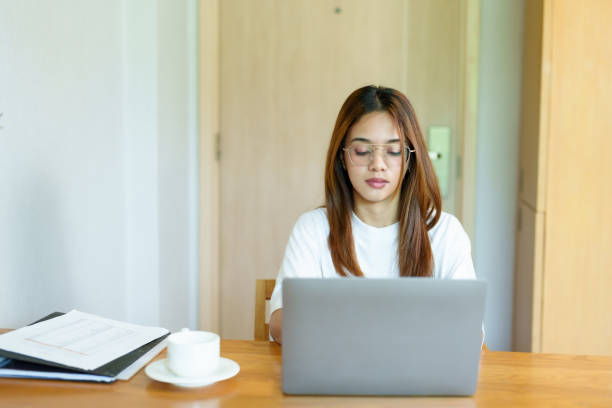 istock Beautiful Asian woman working from home. Closing eyes with a tired expression while working on laptop. Wearing glasses, brown hair, and white shirt. Experiencing fatigue and stress from work pressure. 2196780398