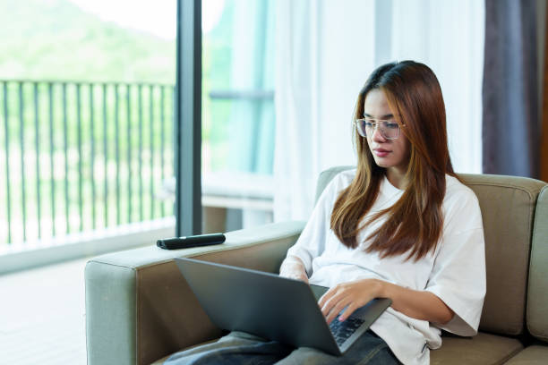 istock Beautiful Asian woman working from home. Sitting on sofa using laptop with serious expression. Large window with natural light, casual white shirt, Remote work lifestyle, modern home setting. 2196780409