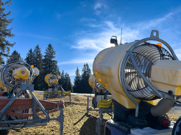 A group of large snow cannons positioned on a mountain slope, ready for artificial snow production. The bright yellow machines stand against a clear blue sky, surrounded by evergreen trees, highlighting the technology used in ski resorts to maintain optimal winter conditions. Perfect for winter sports, ski industry, and climate adaptation visuals.