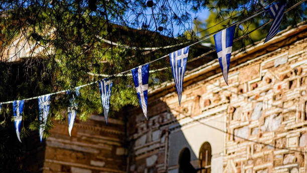 This image features Greek flags strung across the front of a historic church in Athens, framed by lush pine tree branches. The traditional Byzantine architecture in the background highlights the intricate details and cultural significance of the site. The combination of national symbols and historical elements evokes a sense of Greek heritage, tradition, and national pride, set against a serene and vibrant environment.