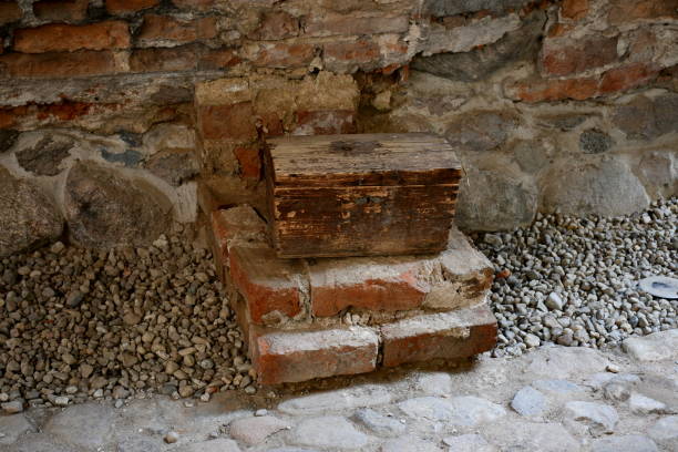A close up on a small chest located on a stand made out of red and grey bricks situated next to a path or road made out of stones, rocks, or boulders, and a sturdy yet damaged wall made out of bricks