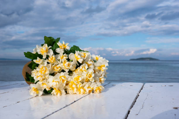 A bouquet of vibrant yellow daffodils rests on a white wooden table, set against a serene sea view. A perfect scene for spring, nature, and tranquility themes.