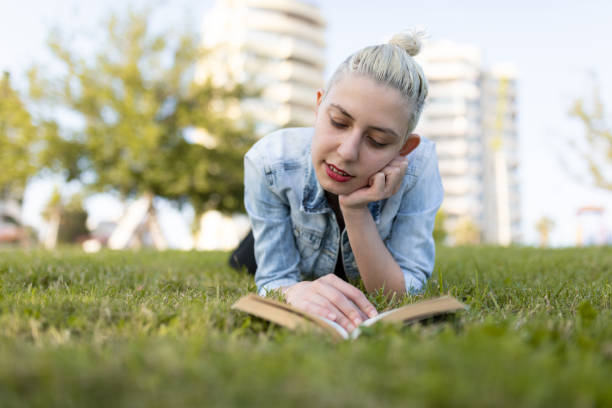 Young woman with blonde hair tied in a bun lying on the grass in a park while reading a book. She is wearing a denim jacket and black pants, with modern apartment buildings and green trees in the background.