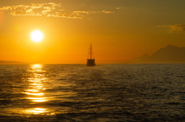 Sailing ship at sunset over a calm sea. Golden light reflects on the water, creating a serene maritime scene with distant mountains in the background.