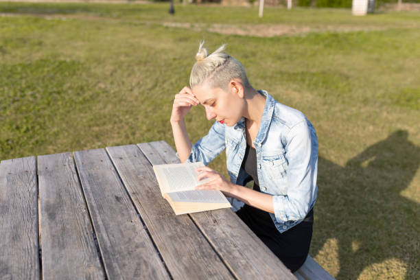 Young woman with short blonde hair tied in a bun sitting on a wooden bench outdoors while reading a book. She is wearing a denim jacket and black pants, with a stone pathway, metal fence, and water in the background.