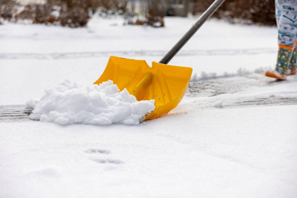 A toddler in rubber boots pushes a yellow snow shovel