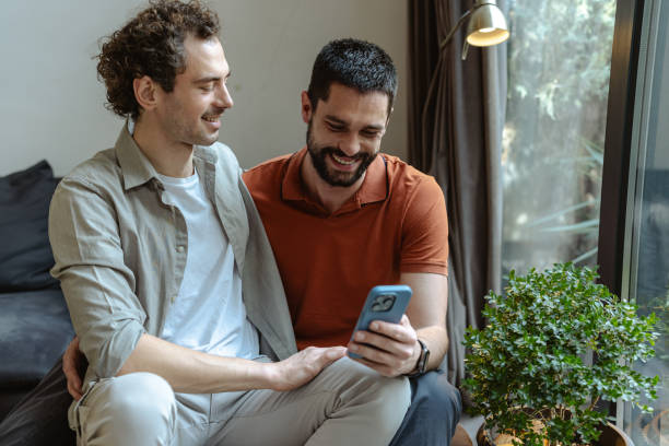 istock Loving gay couple smiling while looking at a smartphone inside a cozy home 2196839843
