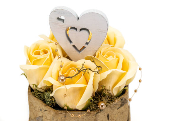 A delicate arrangement of yellow roses accompanied by a wooden heart decoration with intricate cutouts. The composition is enhanced by gold beads and a rustic base, creating a warm and elegant aesthetic.