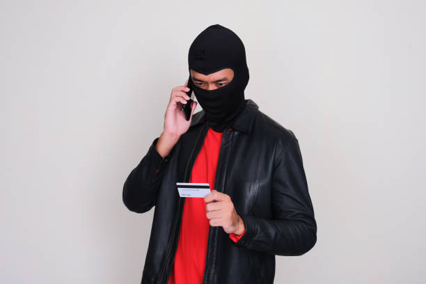 A scammer wearing balaclava holding credit card when calling his victim A scammer wearing balaclava holding credit card when calling his victim telepon uang stock pictures, royalty-free photos & images