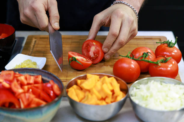 Fresh vegetables are being chopped with a Japanese Bunka knife on a wooden cutting board. Colorful bell peppers, tomatoes, squash, and onions are prepared for a meal.