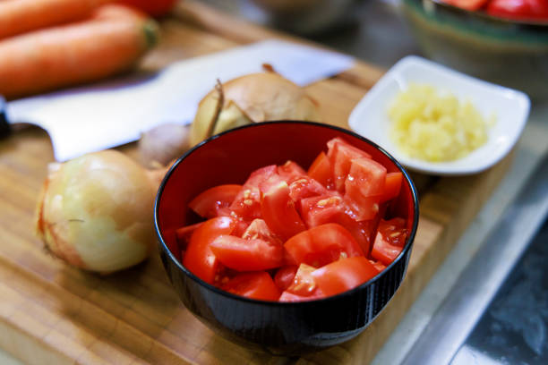 Chopped fresh tomatoes are placed in a black bowl on a wooden cutting board. Nearby are onions, carrots, and small bowls with garlic and other ingredients, ready for cooking.