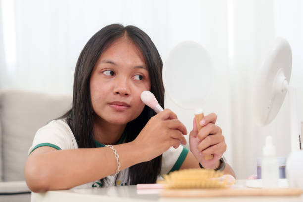 istock Asian woman with dark hair applying foundation using a makeup brush while looking into a handheld mirror. Various beauty and skincare products arranged on the white surface. 2196860684