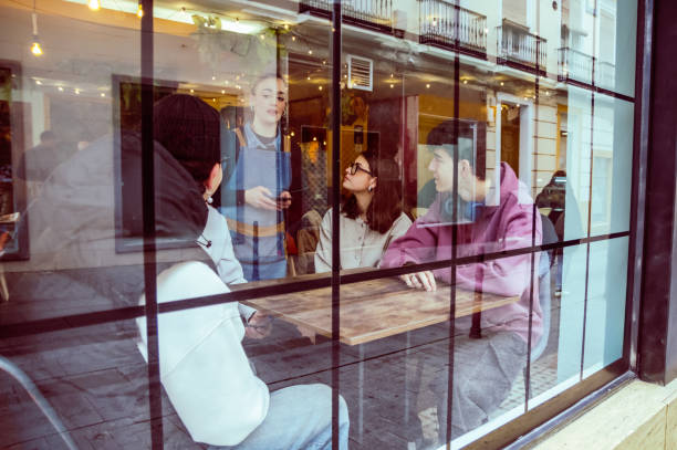 Friendly waitress serving energetic young patrons at bustling cafe table, viewed through window, highlighting lively dining scene