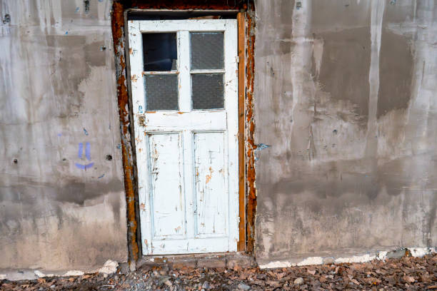 A rustic white wooden door with peeling paint stands against a weathered concrete wall, showcasing signs of aging and time. The cracked texture of the wall and scattered leaves on the ground add to the vintage charm, evoking a sense of abandonment or history. The door’s old glass panels, rusted metal edges, and uneven frame suggest years of exposure to the elements.