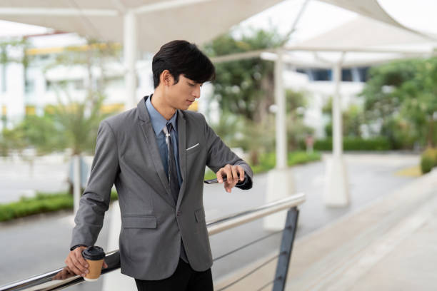 istock Young Asian business professional checking watch and smartphone while holding coffee outside a modern office, representing time management and productivity. 2196895362