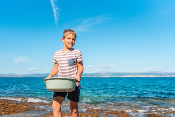 Young Caucasian boy, wearing a striped shirt and shorts, joyfully carries a bucket of freshly caught fish by the vibrant blue sea under a clear sky.