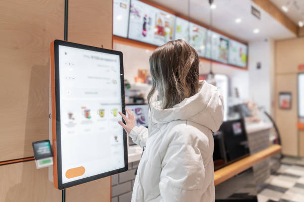 Person Using SelfService Kiosk in Modern Cafe, A woman in a white jacket chooses drinks and orders food for dessert in a restaurant or pizzeria coffee shop through a touchscreen screen on her own. The restaurant menu